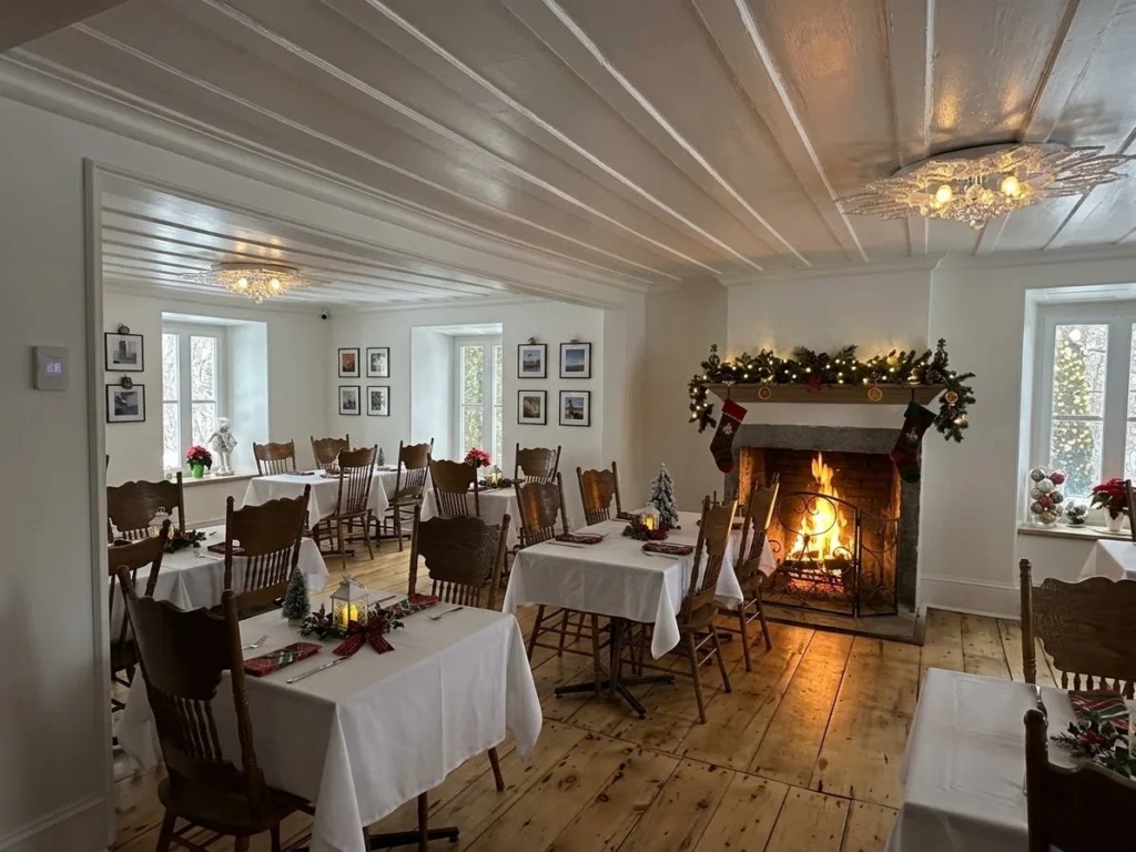 Salle à manger lumineuse de l’Auberge du Manoir avec petites tables et foyer allumé